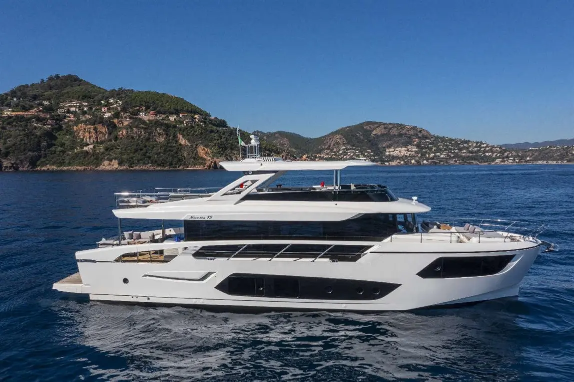 Luxury yacht cruising in clear blue waters near a coastline with green hills and residential areas in the background.